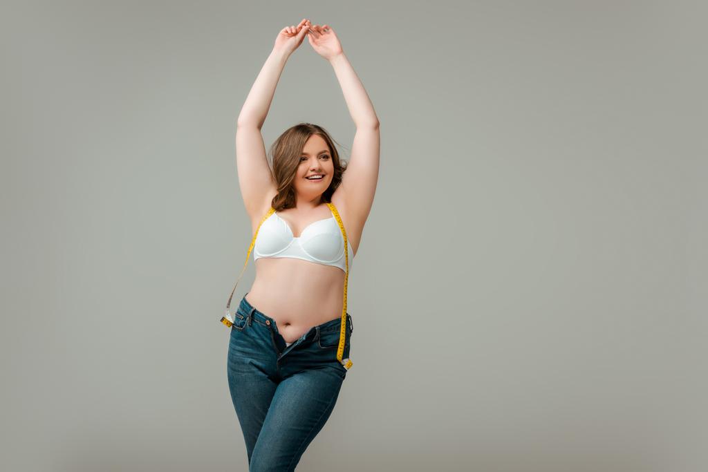 szczęśliwy plus rozmiar kobieta w dżinsach i stanik stojący z rękami nad głową odizolowane na szary - Zdjęcie, obraz