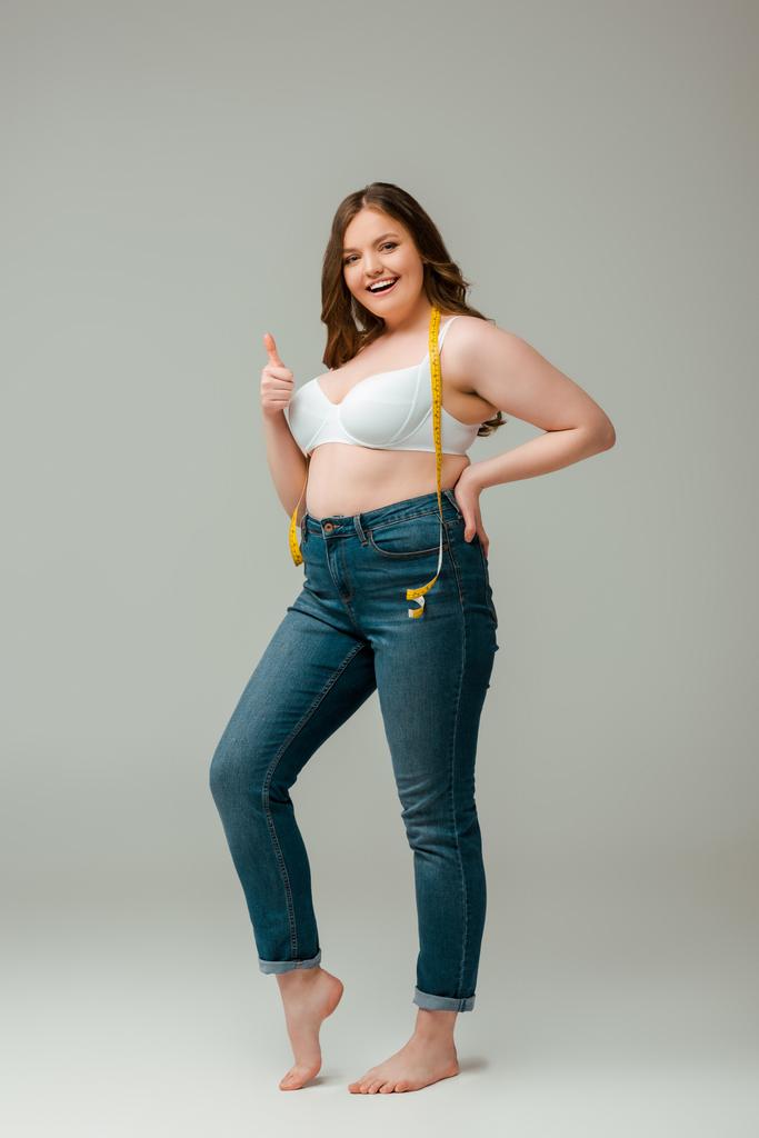 alegre más tamaño mujer en jeans y sujetador de pie con cinta métrica y mostrando el pulgar hacia arriba en gris
 - Foto, Imagen