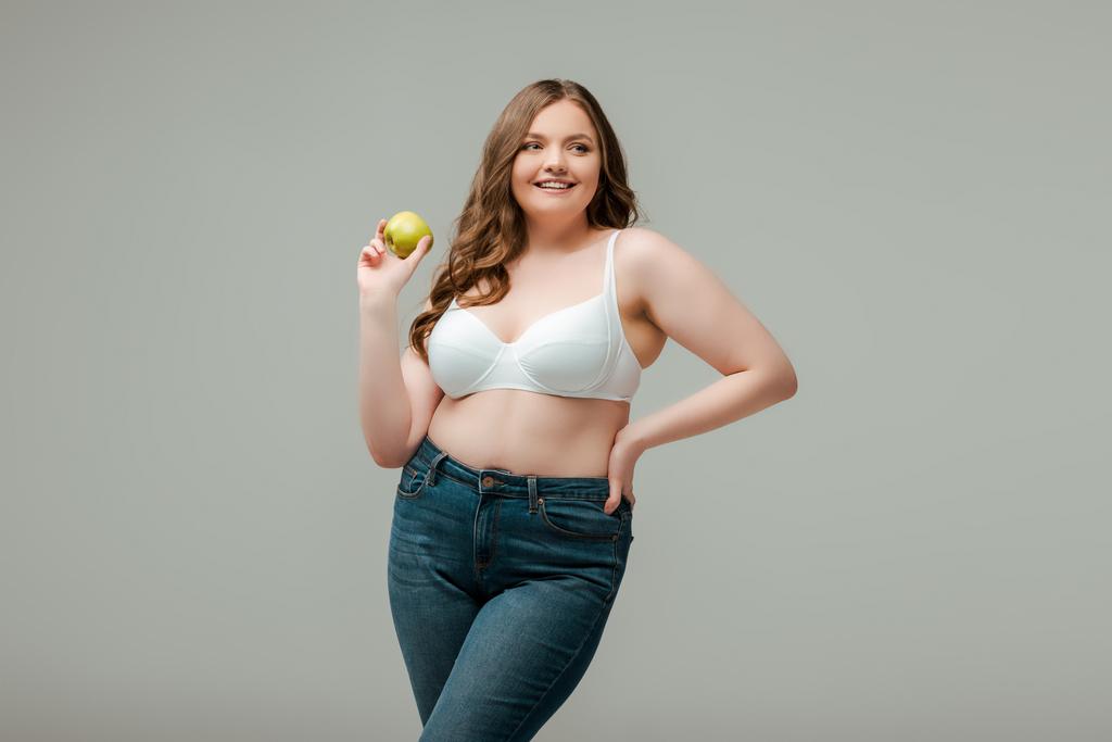 glückliche Plus-Size-Frau in Jeans und BH hält Apfel isoliert auf grau - Foto, Bild
