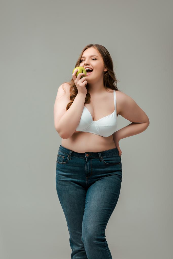 χαρούμενα συν μέγεθος γυναίκα σε τζιν και σουτιέν τρώει μήλο απομονώνονται σε γκρι - Φωτογραφία, εικόνα