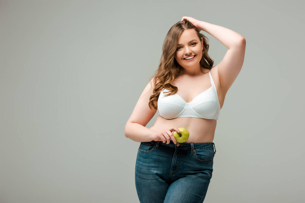 szczęśliwy plus rozmiar dziewczyna w dżinsy i stanik gospodarstwa jabłko izolowane na szary - Zdjęcie, obraz