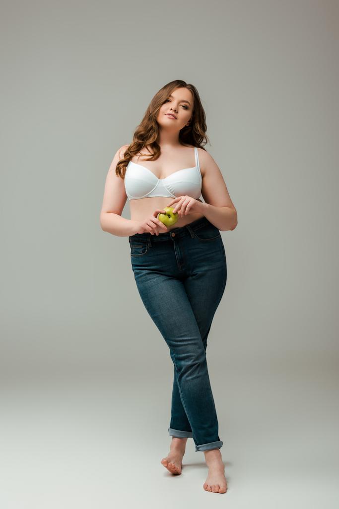 привлекательный плюс размер девушка в джинсах и бюстгальтер держа яблоко на сером
 - Фото, изображение