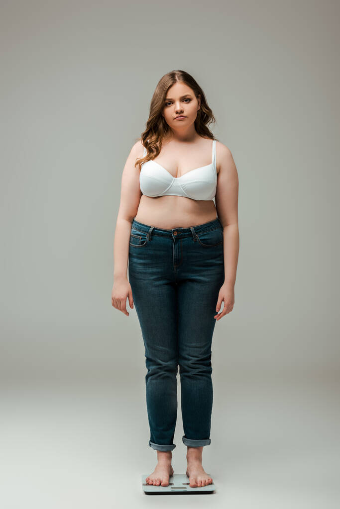 plus size girl in jeans and bra standing on scales and looking at camera on grey - Photo, Image
