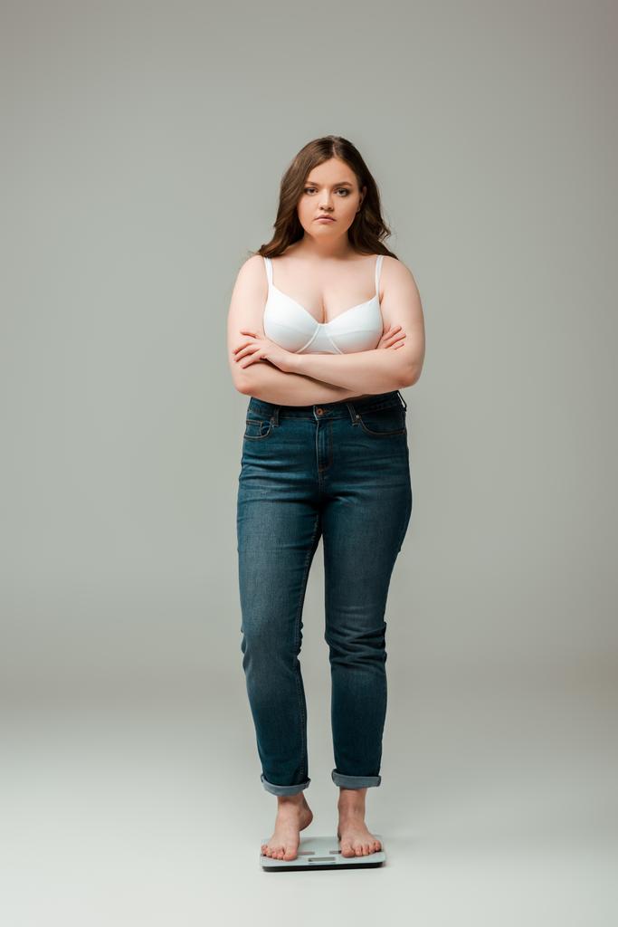 plus velikost dívka v džínách a podprsenka stojící se zkříženými pažemi na váze a při pohledu na kameru na šedé - Fotografie, Obrázek