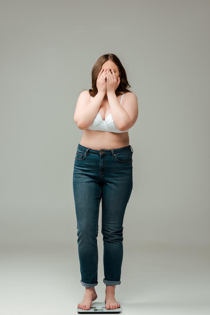 plus size woman in jeans and bra standing on scales and covering face on grey - Photo, Image