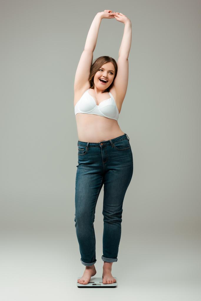 happy plus size woman in jeans and bra standing with hands above head on scales and smiling on grey - Photo, Image