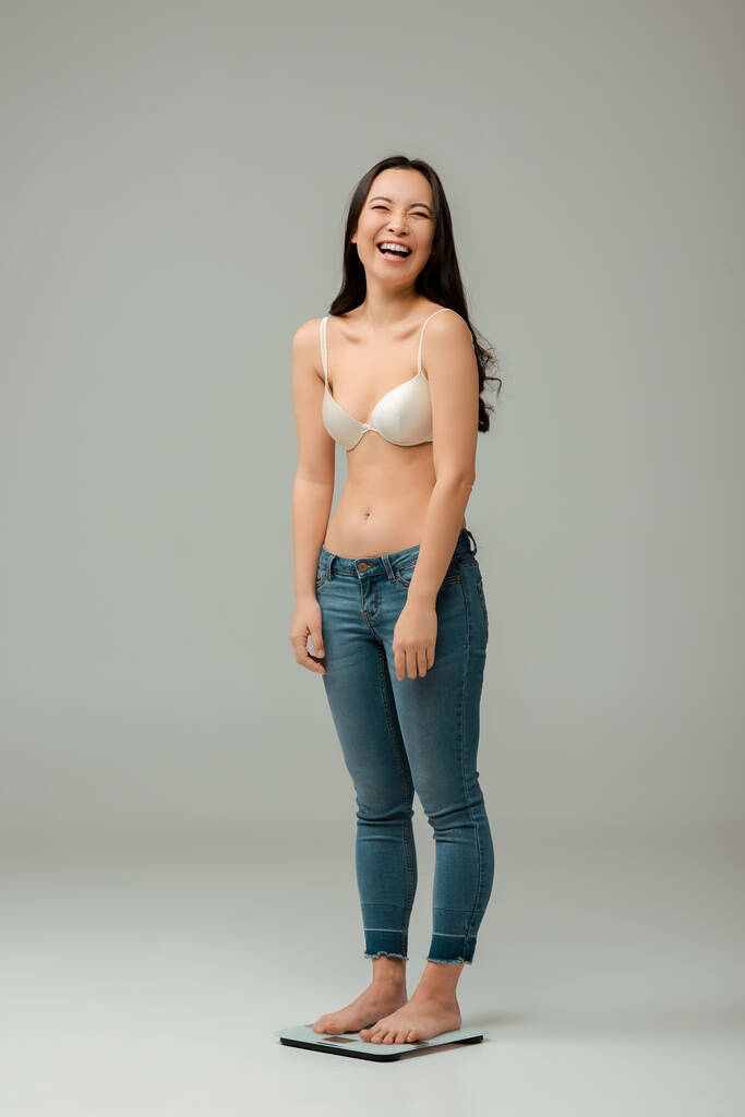 šťastná asijská žena stojící na stupnici a smějící se na šedé - Fotografie, Obrázek