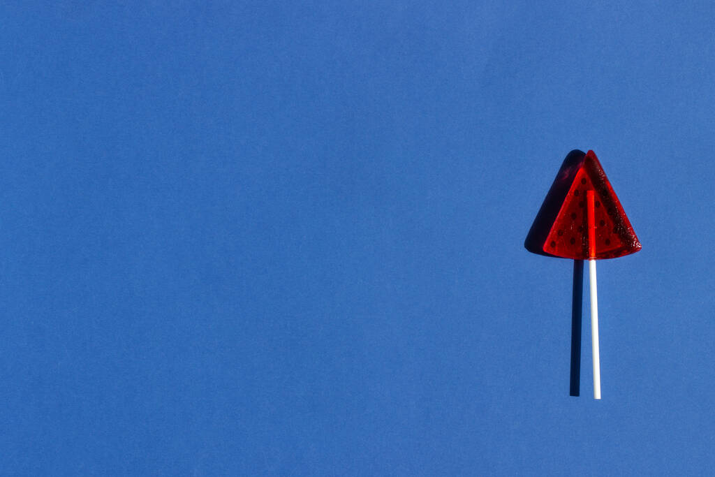 Красный леденец в форме ломтика арбуза на ярко-голубом фоне с ярким солнечным светом и твёрдыми тенями. Летний отдых
. - Фото, изображение