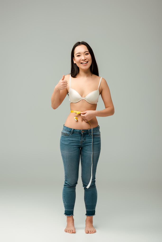 onnellinen ja ylipainoinen aasialainen tyttö rintaliivit mittaus vyötärö ja osoittaa peukalo ylös harmaa
  - Valokuva, kuva