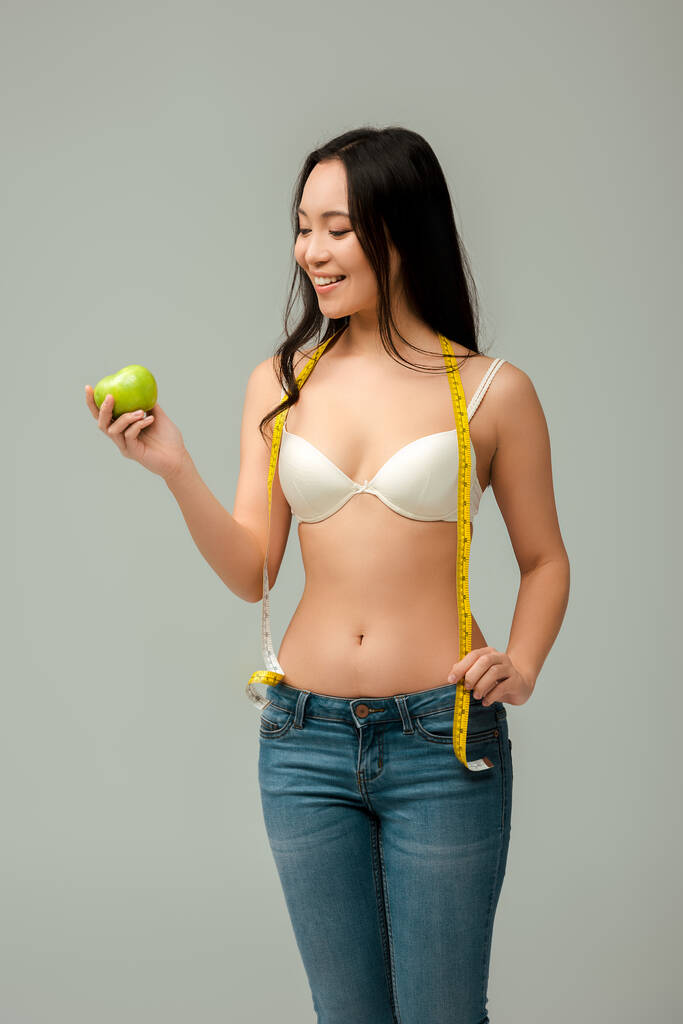 весела і надмірна вага азіатська дівчина тримає мірну стрічку і яблуко ізольовано на сірому
  - Фото, зображення