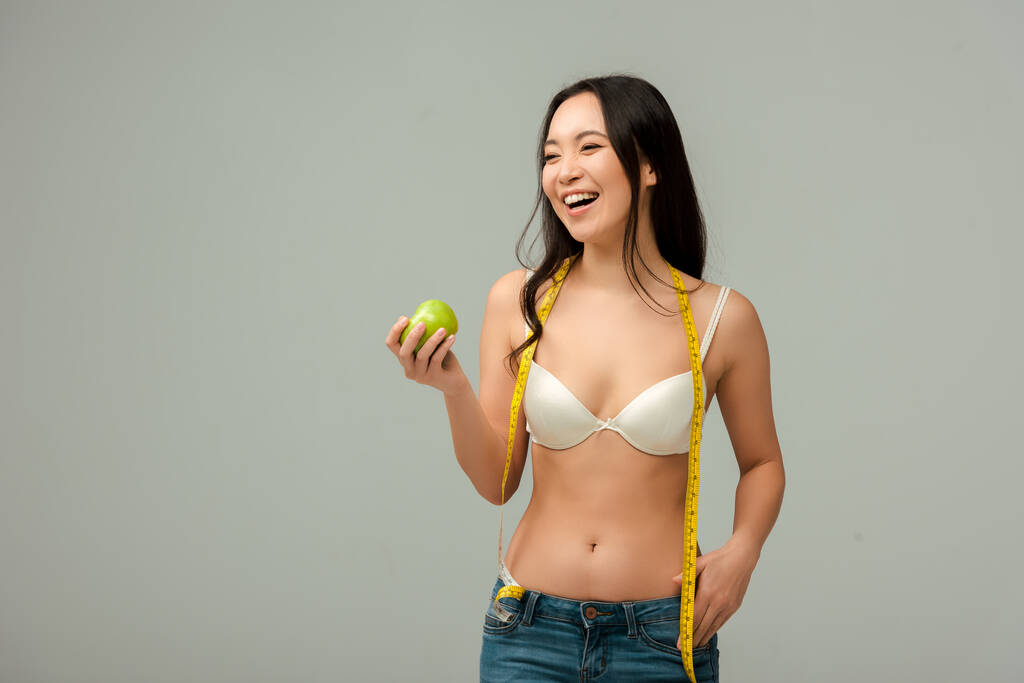 šťastný a nadváha asijské dívka drží měřicí pásku a jablko izolované na šedé  - Fotografie, Obrázek