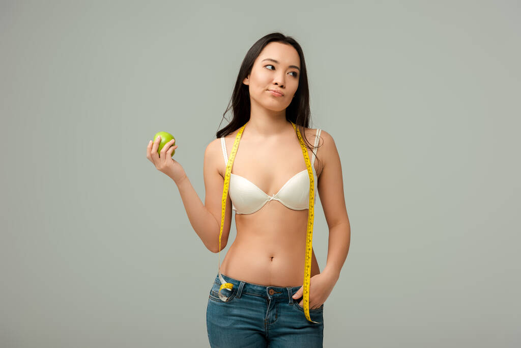 недовольная азиатская девушка держит яблоко, стоя с рукой в кармане изолированы на серый
  - Фото, изображение
