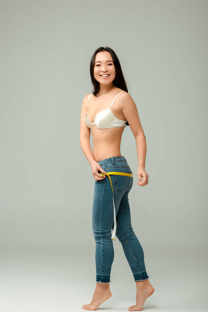 gelukkig en overgewicht aziatisch meisje in beha meten heupen op grijs  - Foto, afbeelding