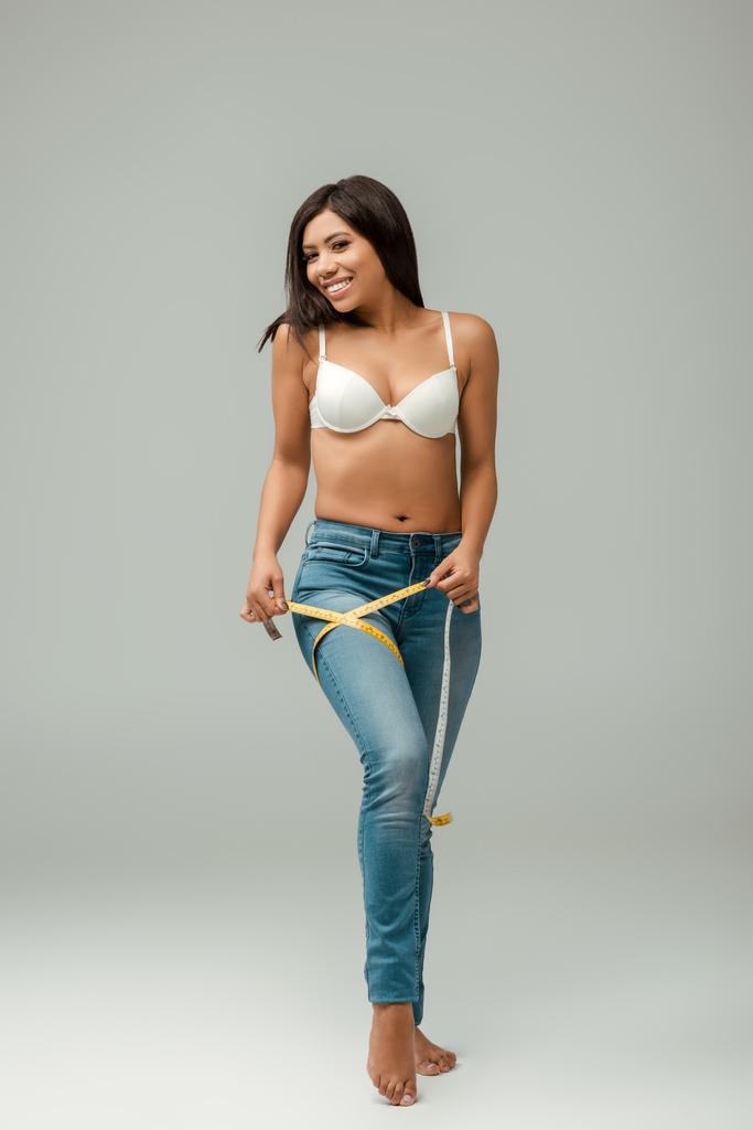 sorridente e sovrappeso ragazza afroamericana in jeans e reggiseno misura coscia su grigio
 - Foto, immagini