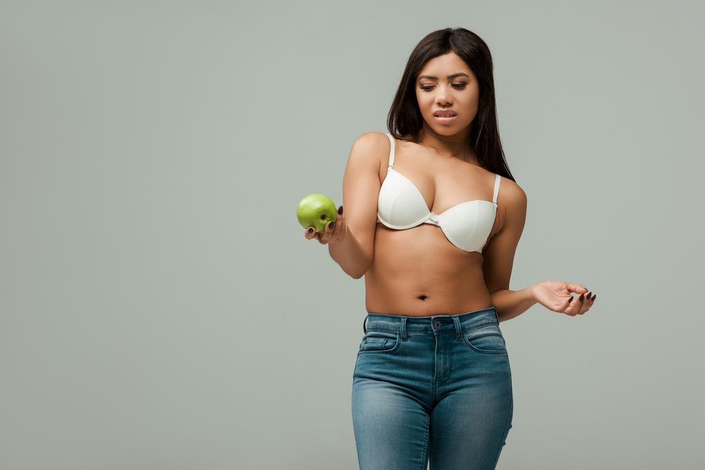 unzufriedene und übergewichtige Afroamerikanerin in Jeans und BH schaut auf Apfel isoliert auf grau - Foto, Bild