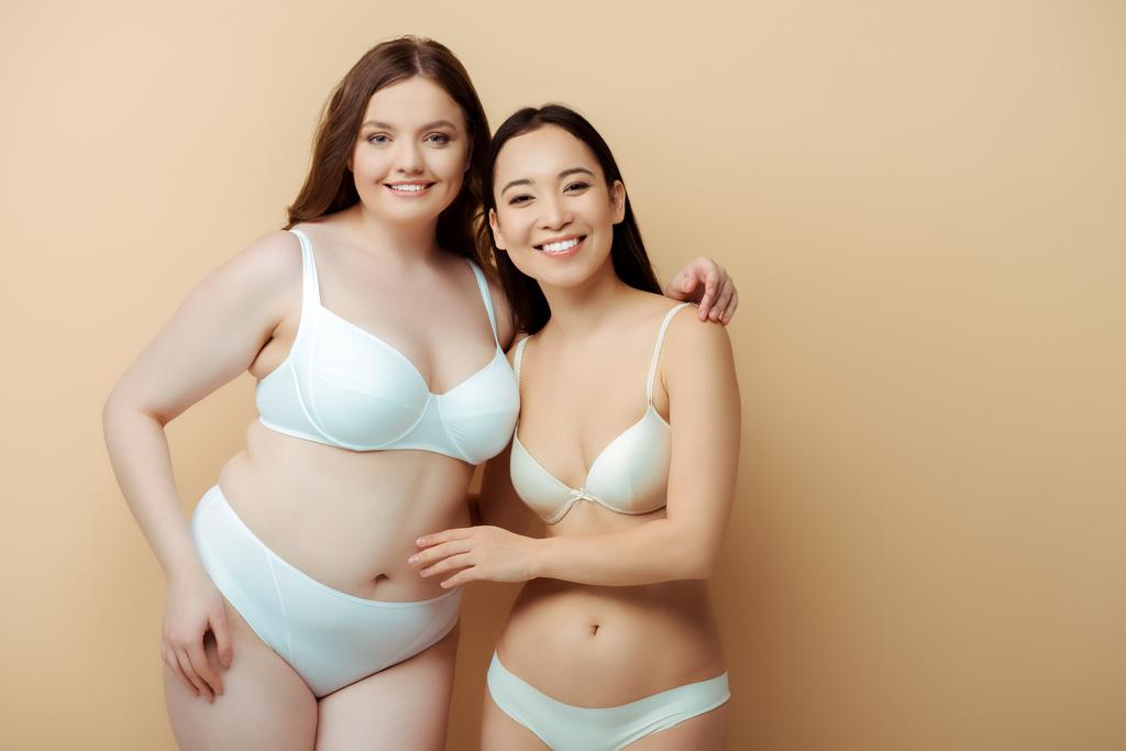 plus size Frau lächelt mit asiatischem Mädchen in Unterwäsche isoliert auf beige, Körper positives Konzept  - Foto, Bild