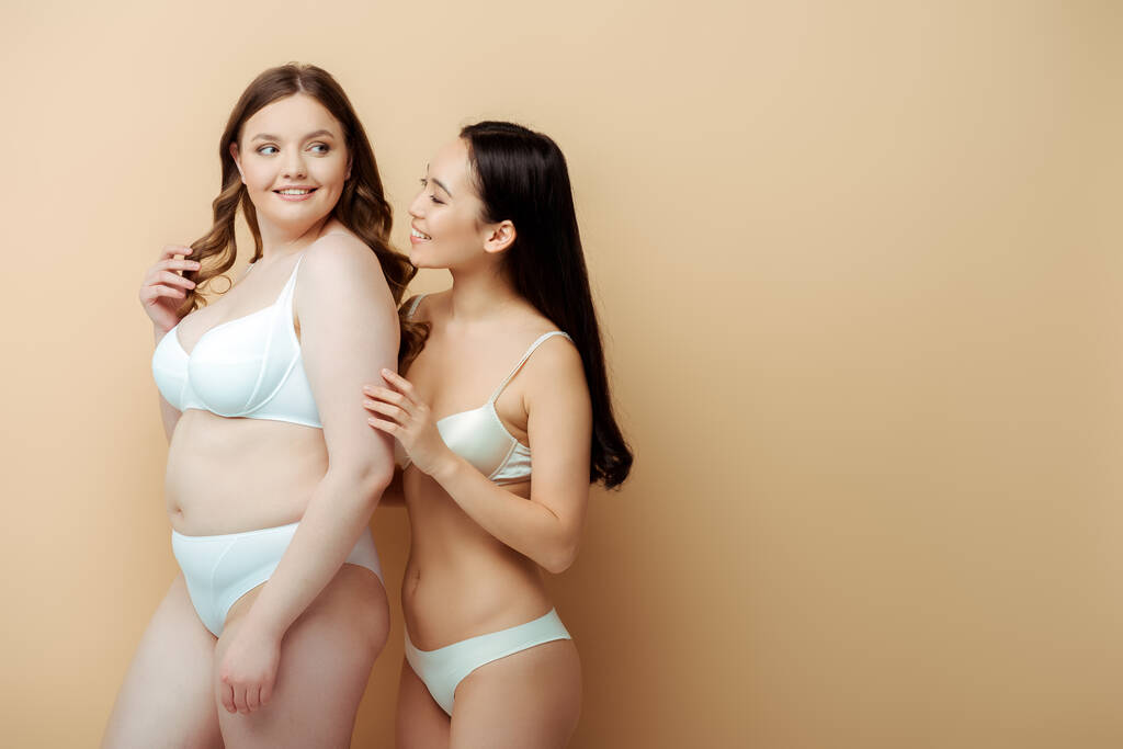 fröhliche Plus-Size-Frau steht mit asiatischem Mädchen in Unterwäsche isoliert auf beige, körperpositives Konzept  - Foto, Bild