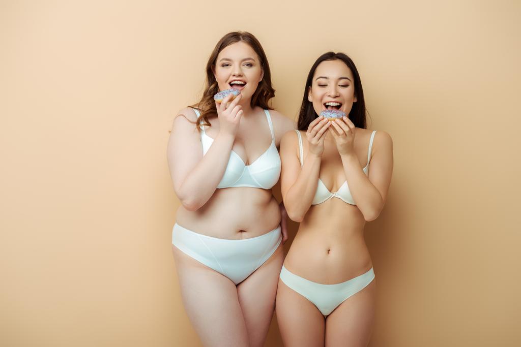 feliz más tamaño mujer y asiático chica en ropa interior comer donas aislado en beige, cuerpo positivo concepto
  - Foto, Imagen