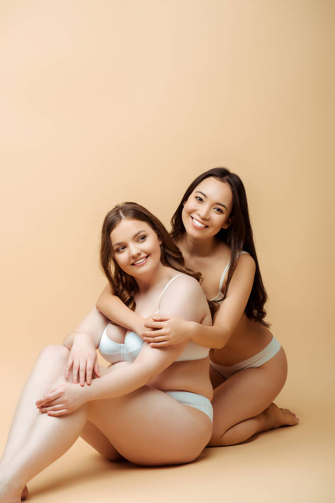 felice ragazza asiatica abbracciando donna in sovrappeso in biancheria intima mentre seduto sul beige, il corpo concetto positivo
  - Foto, immagini