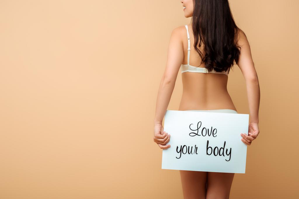 abgeschnittene Ansicht von glücklichen Mädchen mit Plakat mit Liebe deinen Körper Schriftzug isoliert auf beige  - Foto, Bild