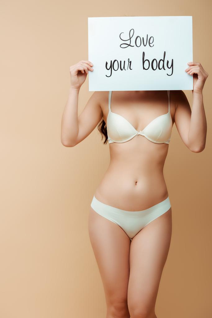 jonge vrouw bedekken gezicht terwijl het houden van plakkaat met liefde uw lichaam belettering geïsoleerd op beige  - Foto, afbeelding