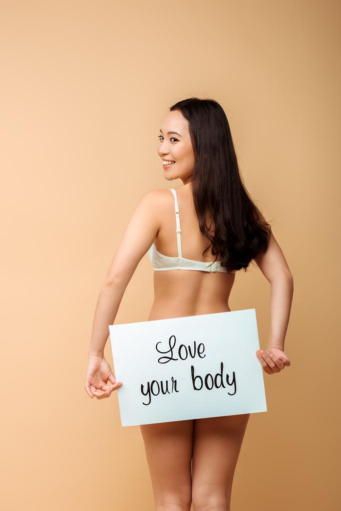 radosna i azjatycka kobieta trzymająca plakietkę z miłością twoje ciało odizolowane na beżu  - Zdjęcie, obraz