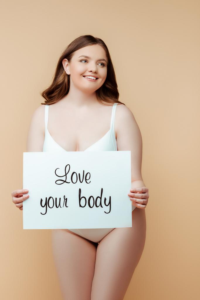 cheerful plus size woman holding placard with love your body lettering isolated on beige, body positive concept  - Photo, Image