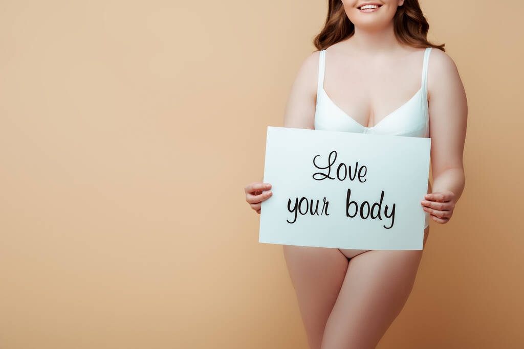 Oříznutý pohled na ženu usmívající se a prezentující plakát s láskou vaše tělo nápisy na béžové pozadí - Fotografie, Obrázek