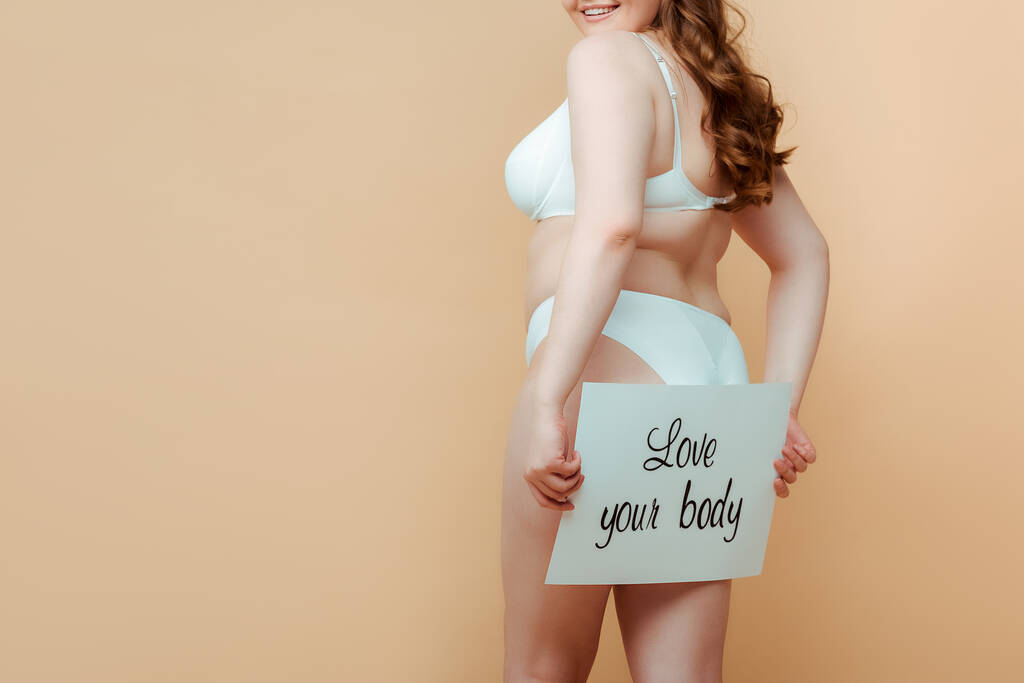 Przycięty widok plus size kobieta prezentując tabliczkę z miłości twoje ciało napis izolowany na beżu - Zdjęcie, obraz