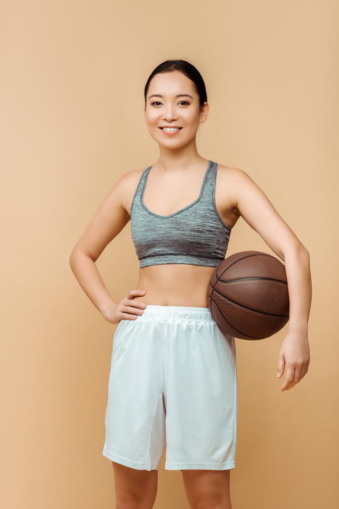 Femme sportive asiatique avec la main sur la hanche et la balle souriant et regardant la caméra isolée sur beige
 - Photo, image