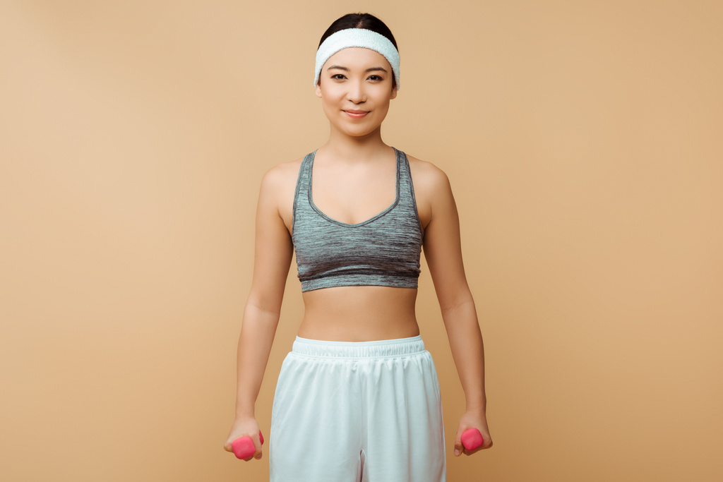 Asian sportswoman with dumbbells smiling and looking at camera on isolated on beige - Photo, Image