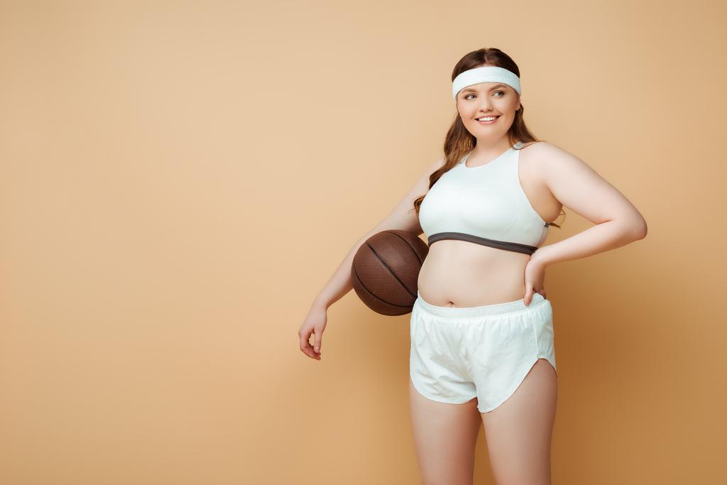 Plus size sportswoman with hand on hip and ball smiling and looking away on beige - Photo, Image