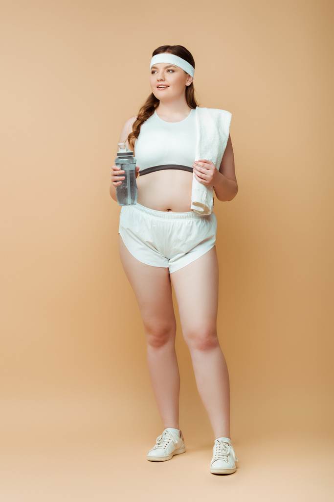 Plus size sportswoman with towel and sports bottle looking away on beige - Photo, Image