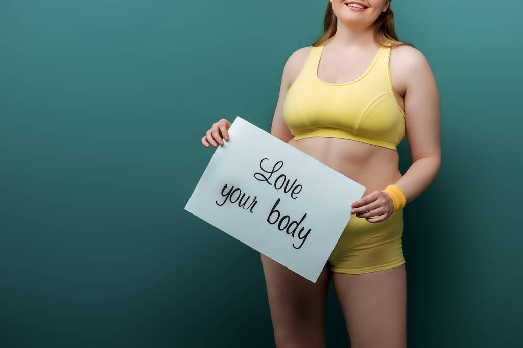 Ausgeschnittene Ansicht einer Plus-Size-Sportlerin, die lächelt und ein Plakat mit Love your body Schriftzug auf Grün zeigt - Foto, Bild