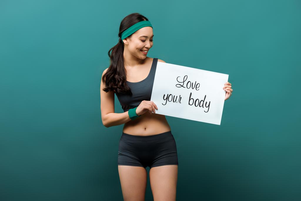 Front view of asian sportswoman smiling and showing placard with love your body lettering on green background - Photo, Image