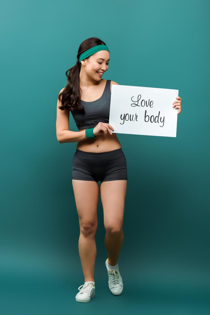 Привлекательная азиатская спортсменка улыбается и смотрит на плакат с любовью ваше тело надпись на зеленом
 - Фото, изображение