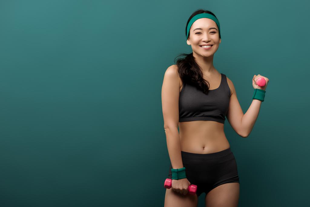 Asiatische Sportlerin lächelt, blickt in die Kamera und trainiert mit auf Grün isolierten Hanteln - Foto, Bild