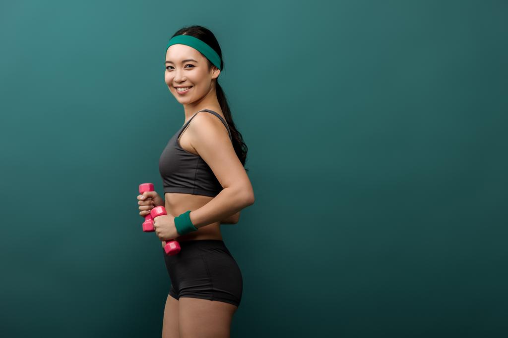 Seitenansicht einer asiatischen Sportlerin mit Hanteln, die lächelt und isoliert auf Grün in die Kamera blickt - Foto, Bild