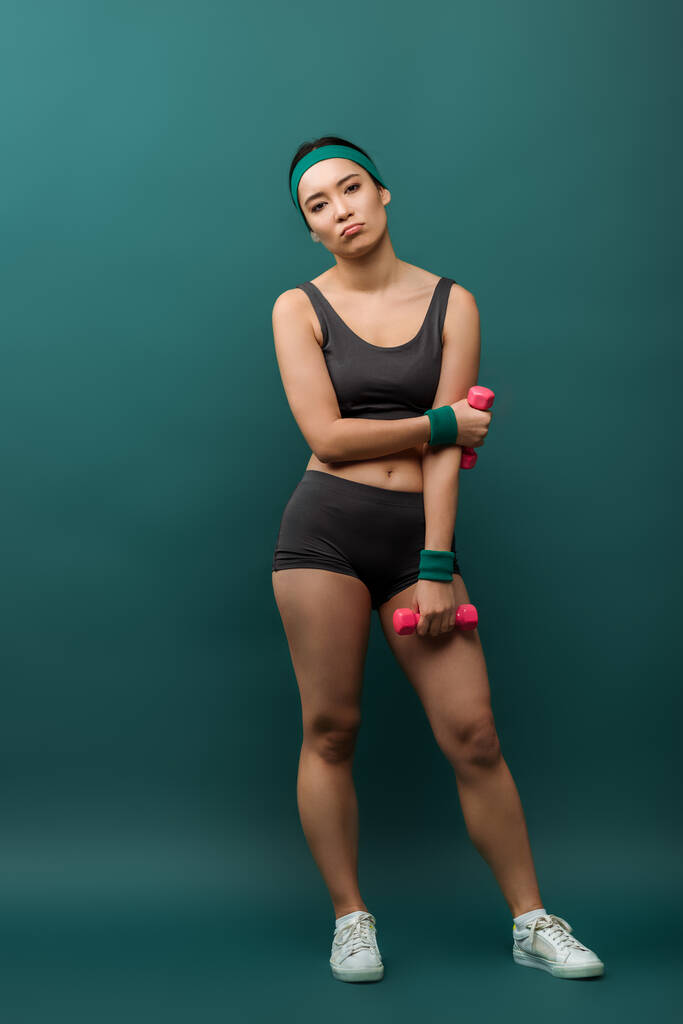 Moe aziatische sportvrouw met halters op zoek naar camera op groene achtergrond - Foto, afbeelding