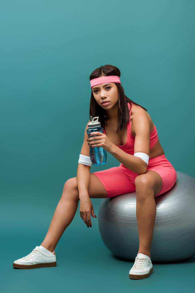 Attraktive afrikanisch-amerikanische Sportlerin hält Sportflasche in der Hand und schaut in die Kamera auf Fitnessball auf grünem Hintergrund - Foto, Bild
