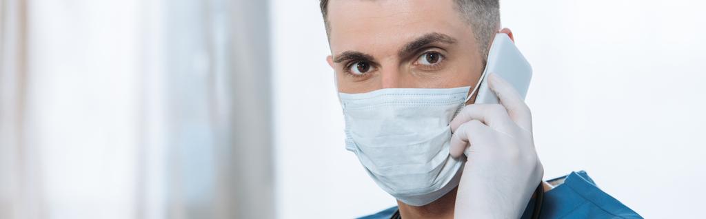 panoramic shot of young doctor in medical mask and latex gloves talking on smartphone and looking at camera - Photo, Image