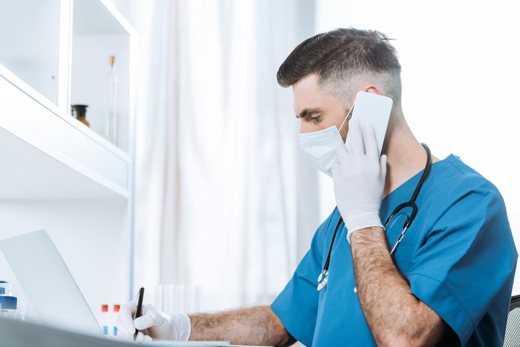 selective focus of young doctor in medical mask and latex gloves writing while talking on smartphone - Photo, Image