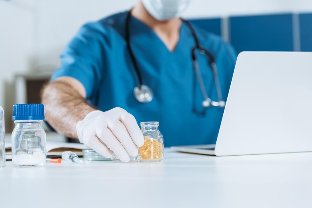 vista recortada do médico em luvas de látex tomando recipiente de vidro com cápsulas enquanto sentado perto do laptop
 - Foto, Imagem