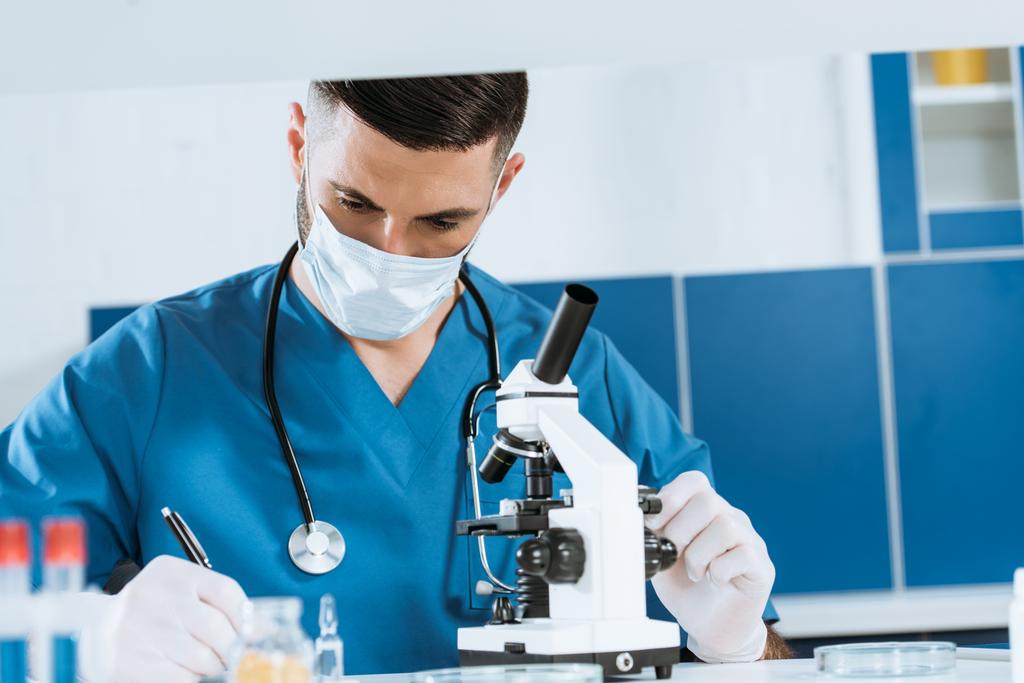 Tıbbi maskeli ve lateks eldivenli genç biyologlar laboratuarda mikroskoba yakın yazıyorlar. - Fotoğraf, Görsel