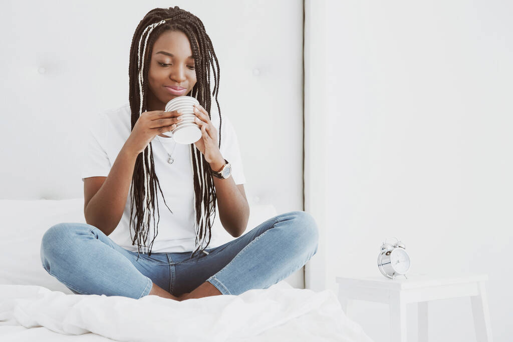 A beautiful African woman sits in a lotus pose on a bed in a white bedroom and drinks hot coffee, an hour from a cup. The girl is dressed in tight jeans and a T-shirt, pigtails on her head. - Photo, Image
