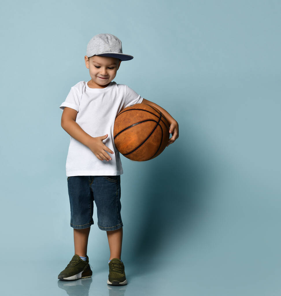 Niño pequeño en camiseta blanca, gorra gris, pantalones cortos de mezclilla, zapatillas de deporte de color caqui. Sosteniendo la pelota de baloncesto, sonriendo, posando sobre fondo azul
 - Foto, Imagen