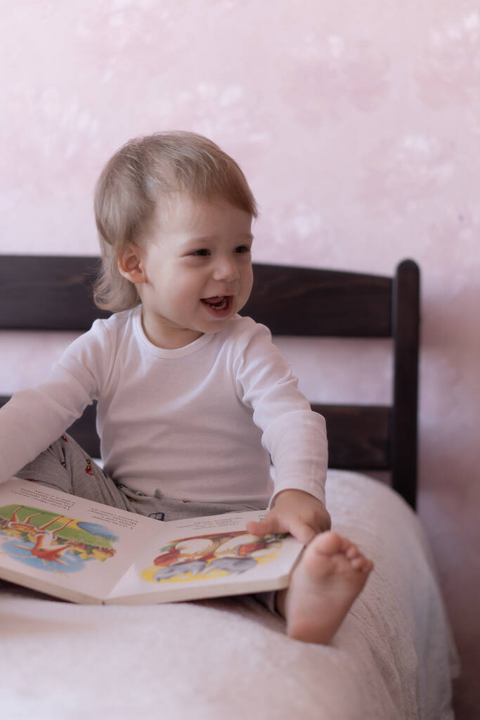 Ein kleiner Junge mit blonden Haaren sitzt auf dem Bett und lächelt, ein Buch in der Hand. Ein kleiner Junge sitzt während der Quarantäne mit einem Buch in der Hand auf einem Bett. - Foto, Bild