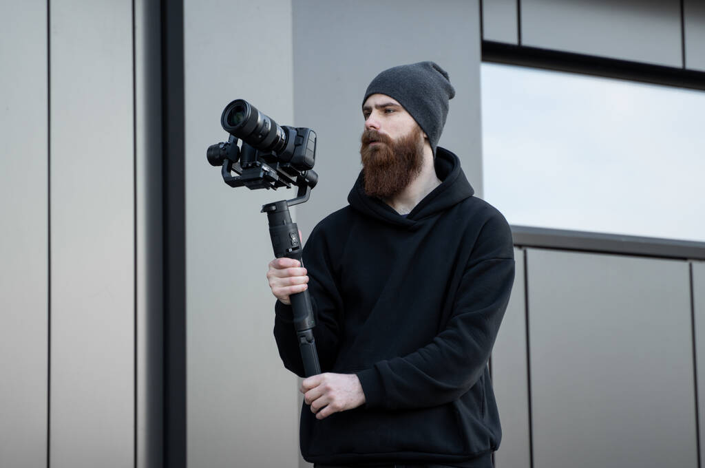 Γενειάδα επαγγελματική videographer σε μαύρο hoodie κρατώντας επαγγελματική κάμερα σε 3-άξονα gimbal σταθεροποιητή. Κινηματογραφιστής κάνει ένα μεγάλο βίντεο με μια επαγγελματική κάμερα κινηματογράφου. Κινηματογράφος. - Φωτογραφία, εικόνα