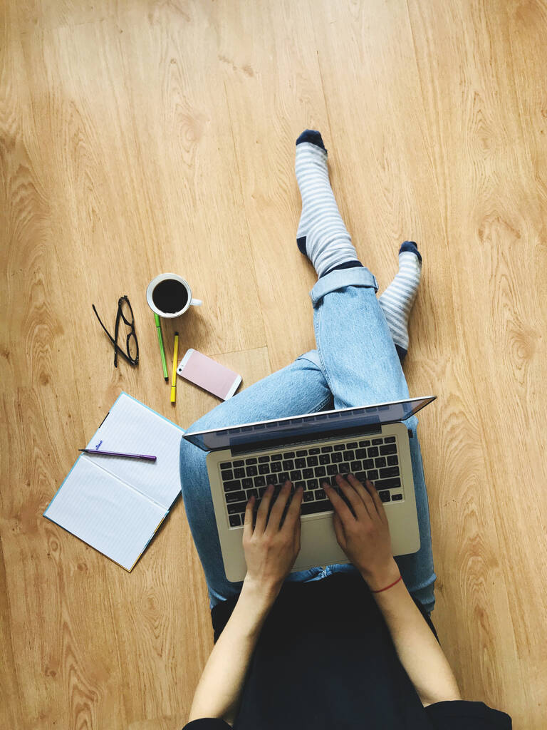 Το κορίτσι που κάθεται στο πάτωμα κρατώντας ένα λάπτοπ στα πόδια της. Σε κοντινή απόσταση βρίσκεται ένα σημειωματάριο, στυλό, τηλέφωνο και μια κούπα καφέ - Φωτογραφία, εικόνα