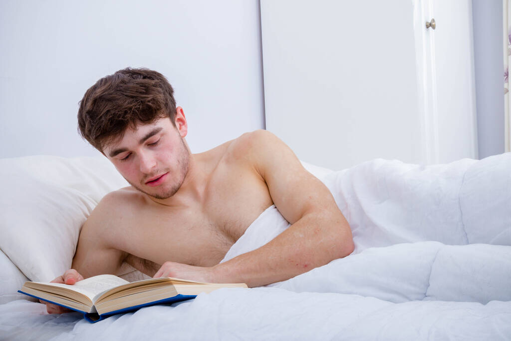 Ein junger erwachsener Mann mit nacktem Oberkörper liest im Bett ein Buch - Foto, Bild